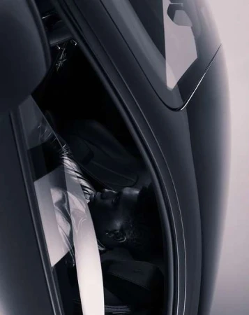 A person lies inside a sleek, dark vehicle, likely a car, viewed at an angle to show the vehicle's interior and the person's resting pose.