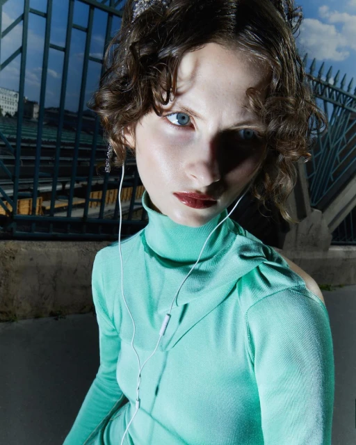 A young woman with curly hair and blue eyes poses outdoors in a light green turtleneck sweater, wearing a tiara and earbuds, against a fence.
