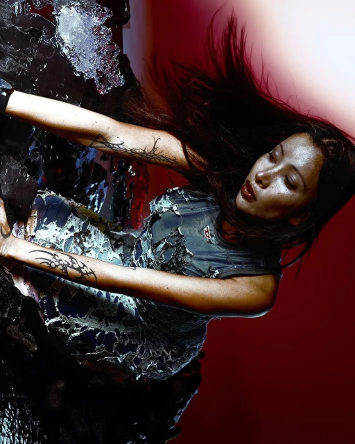 Woman with long dark hair poses in a textured dress with a red background, artistic and abstract, with dramatic lighting creating a striking portrait.