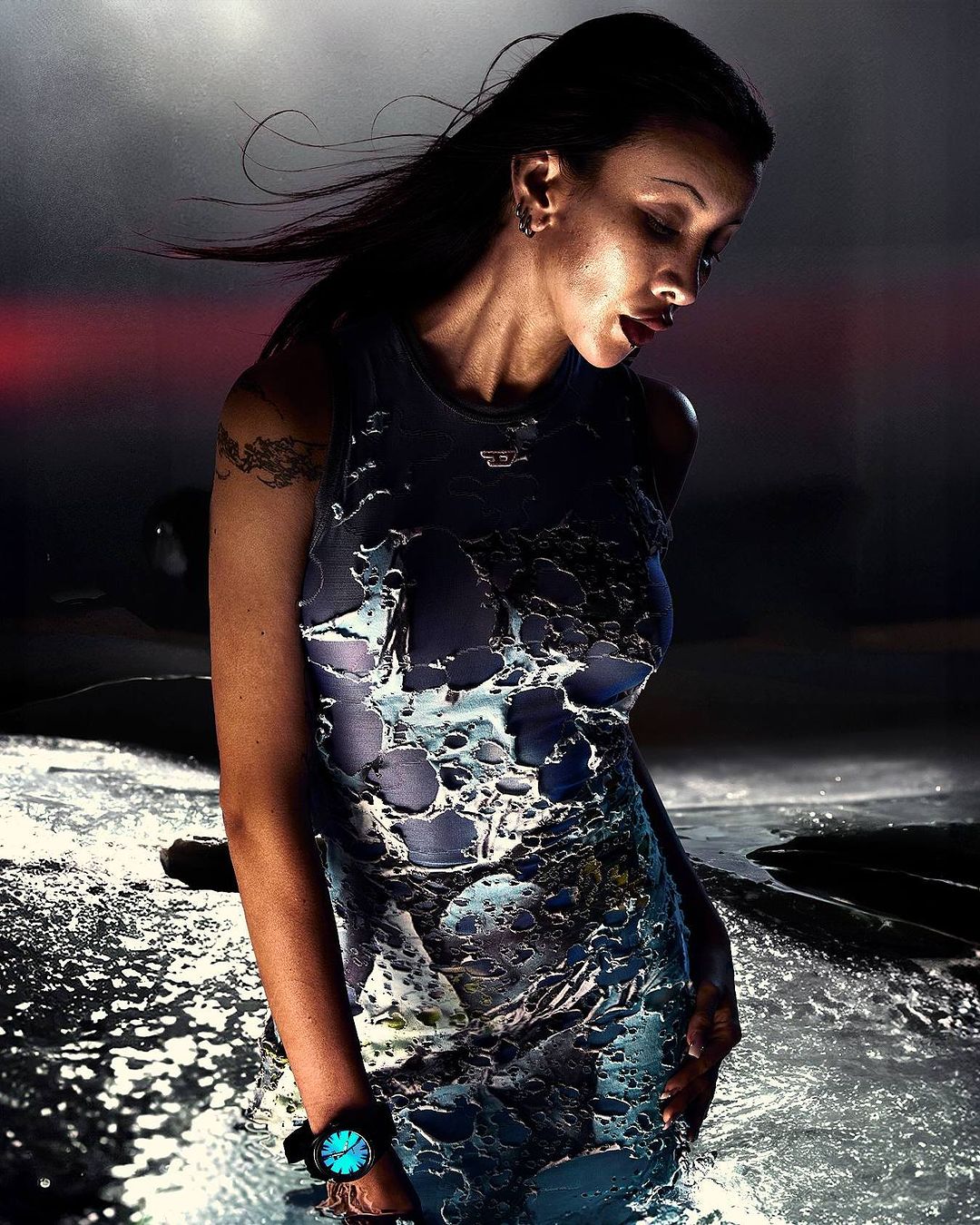 Woman in a form-fitting blue dress stands in a shallow body of water under dark, moody lighting with a red glow in the background.