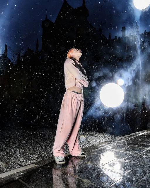 Woman in pink tracksuit stands in rain at night with a dark castle backdrop and glowing orb lights creating an atmospheric scene.