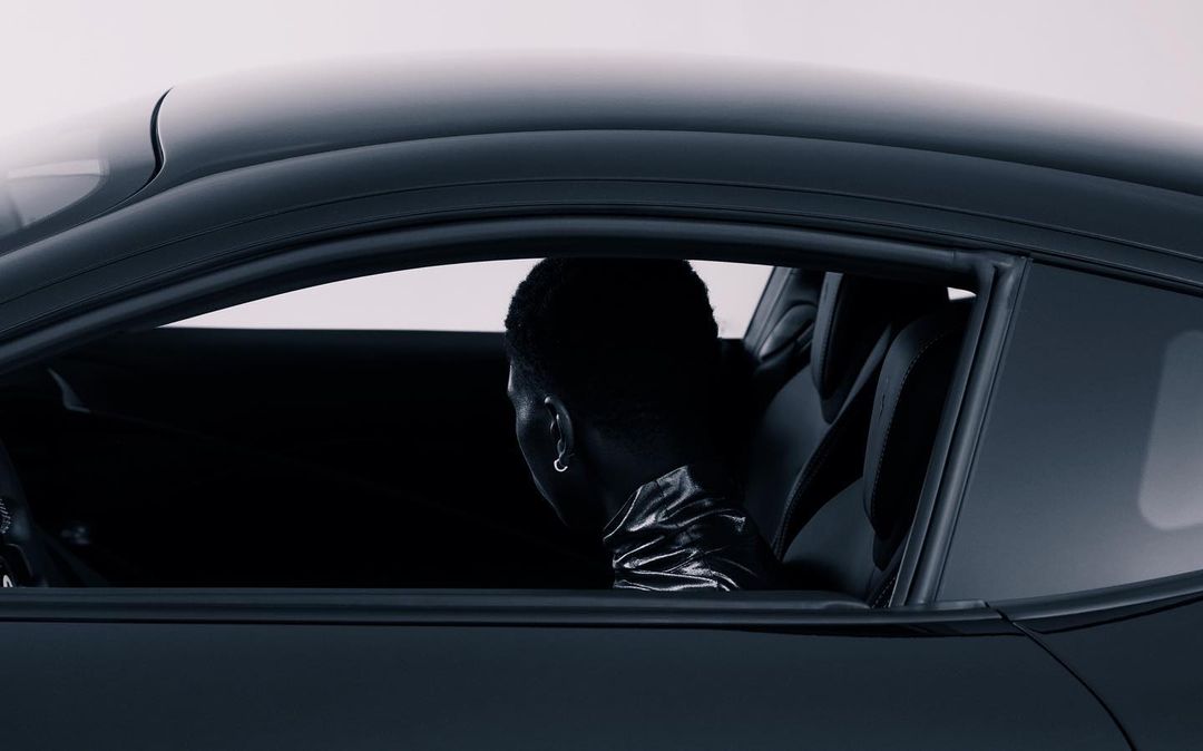 Person inside a black coupe car, viewed from outside. Car interior is visible. Dark aesthetic with bright white background.