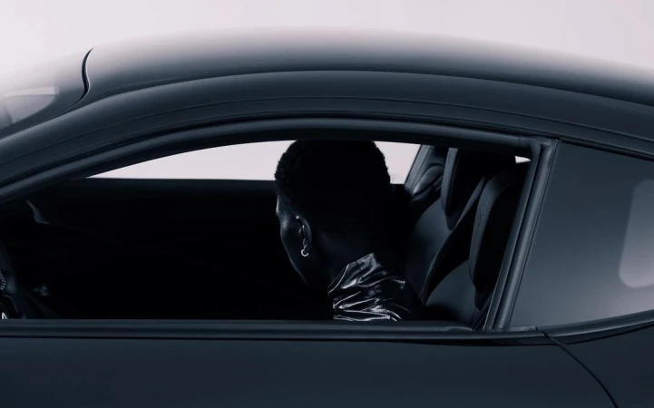 Person inside a black coupe car, viewed from outside. Car interior is visible. Dark aesthetic with bright white background.