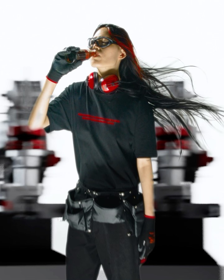 An adult woman drinking from a bottle, with headphones around her neck, wearing a black t-shirt, black gloves, and a tool belt.
