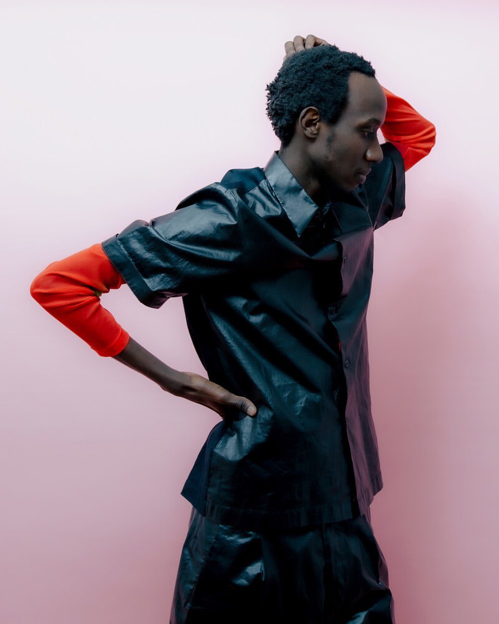 Profile shot of an adult male with dark hair wearing a black collared shirt over a red long-sleeve shirt, posing against a pink background.