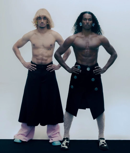 Two muscular shirtless men in contemporary skirts, one with a blonde wig and the other with long dark curly hair, hands on hips.