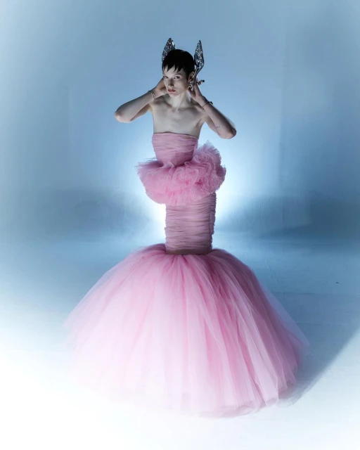 Androgynous fashion model in a pink tulle gown with silver headpiece poses in a photo studio with soft blue light. Beauty and fashion.