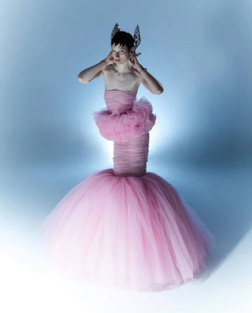 Woman in a pink tulle gown with decorative headpiece poses against a gradient blue background in a fashion photography studio setting.