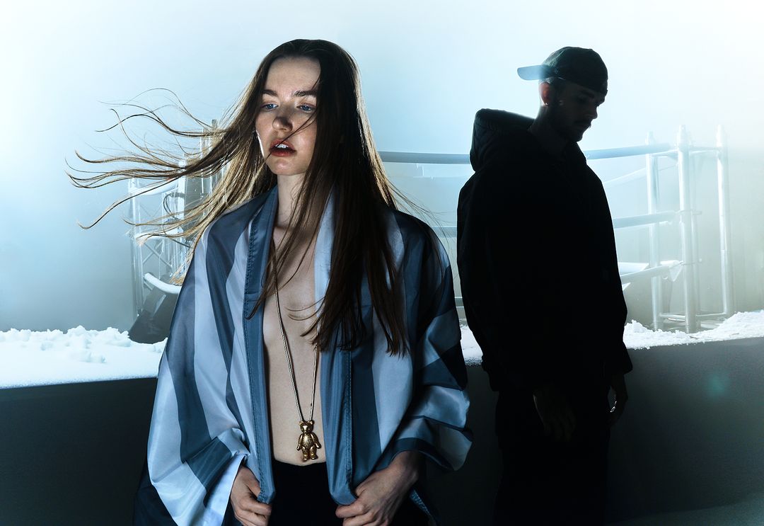 A young woman in a striped jacket and necklace stands indoors while a silhouetted man wearing a baseball cap stands behind her.