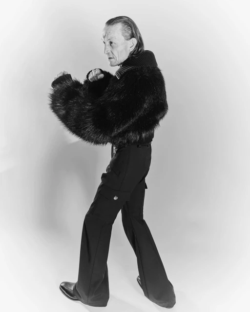 Side view of an older adult posing indoors with fists up, wearing a black faux fur jacket, black pants and shoes against a plain background.