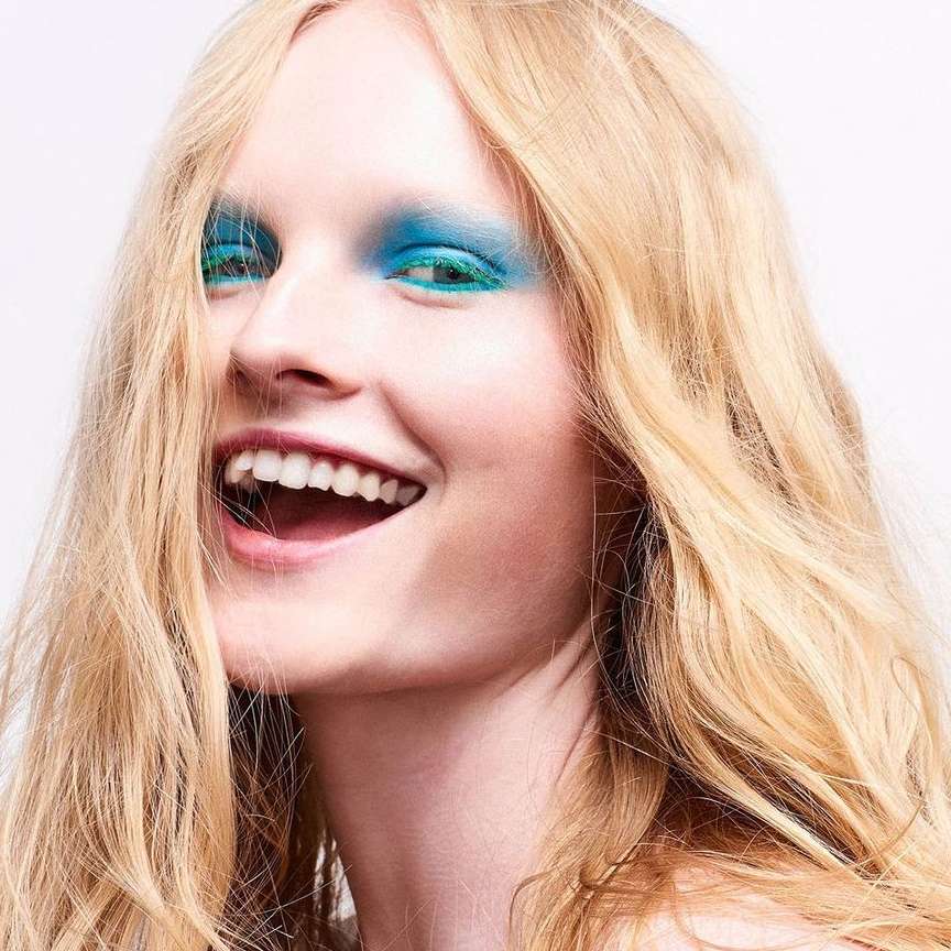 Close-up of a smiling woman with long blonde hair and bright blue eyeshadow, against a plain white background in an indoor setting.