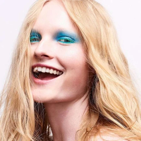 Close-up of a smiling woman with long blonde hair and bright blue eyeshadow, against a plain white background in an indoor setting.