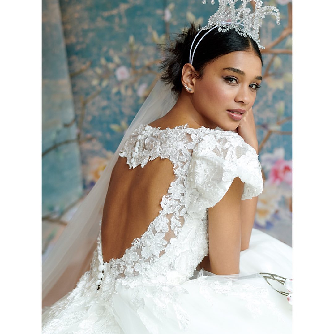 Elegant bride poses in white lace gown and tiara against a blue floral backdrop, showcasing a refined and classic bridal look.