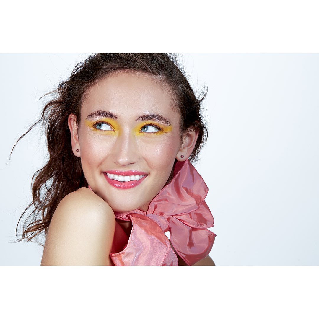 Smiling woman with yellow eyeshadow and a large pink bow. She is indoors against a white background in a beauty or fashion context.
