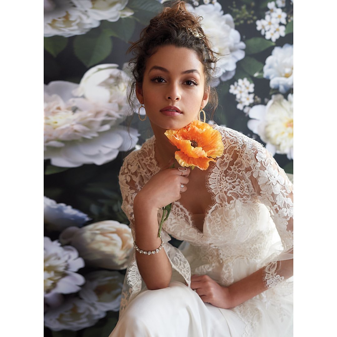 Portrait of a woman in a white lace dress holding an orange flower, set against a backdrop of white roses. Beauty and fashion theme.