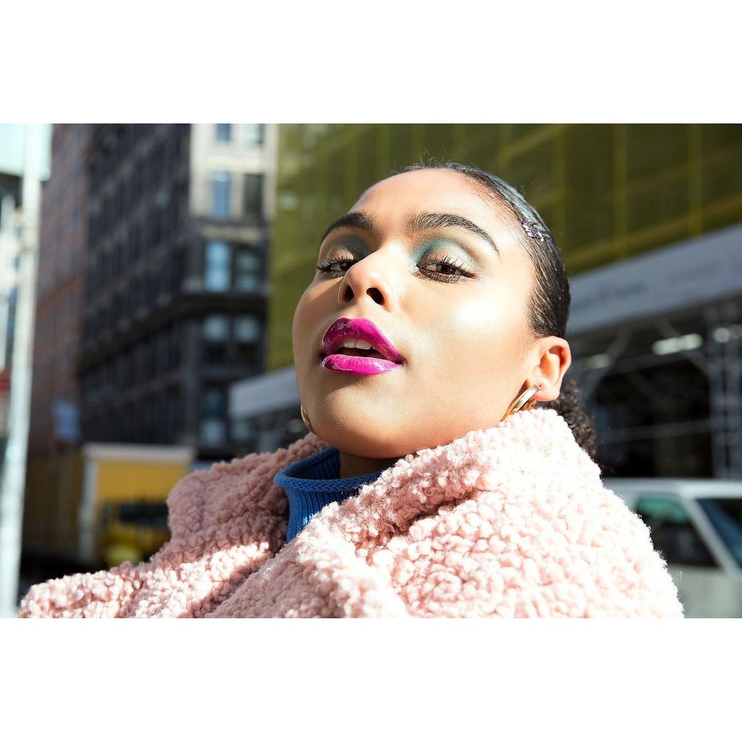Portrait of a young woman with vibrant makeup and a textured pink coat in an urban setting with modern buildings in the background.