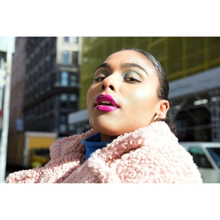 Portrait of a young woman with vibrant makeup and a textured pink coat in an urban setting with modern buildings in the background.