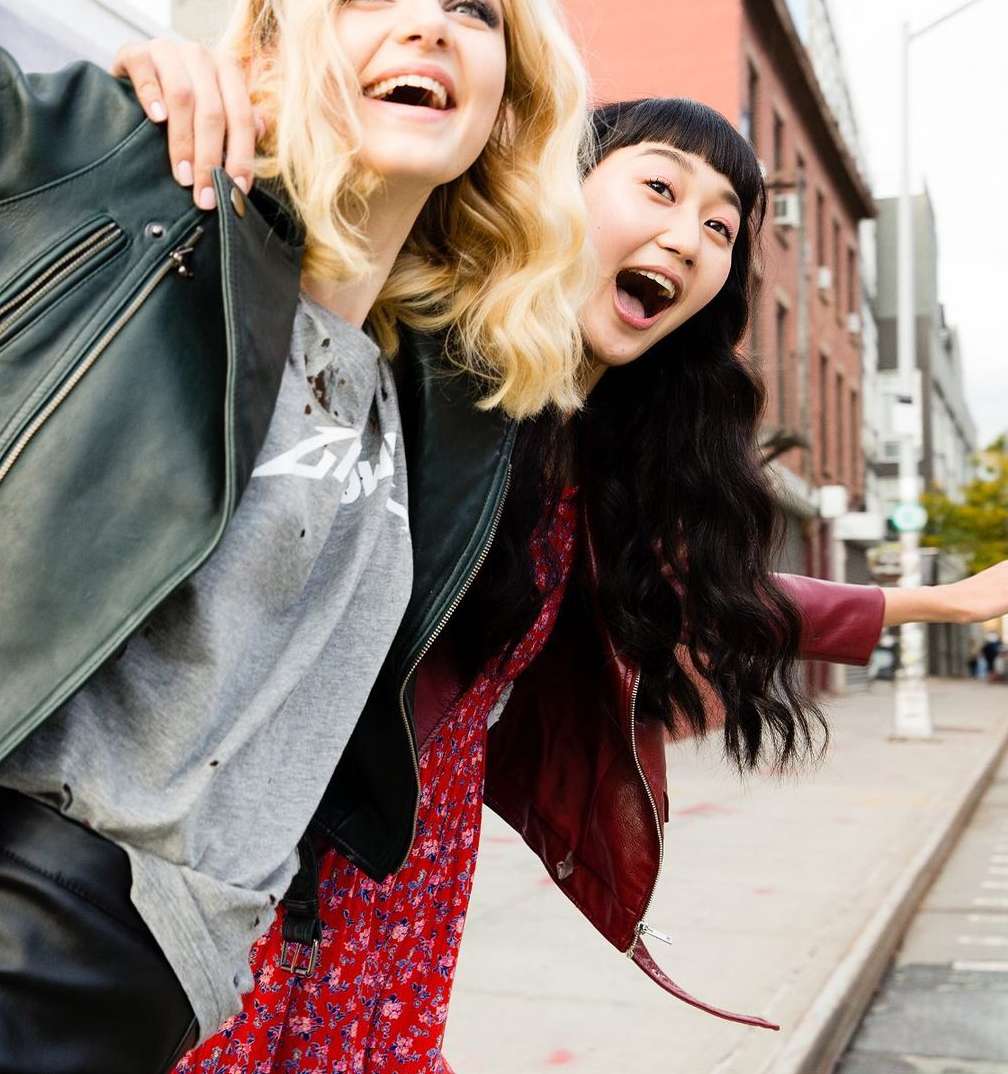 Two women in leather jackets smile on a city street with brick buildings in the background, captured in a medium outdoor shot.