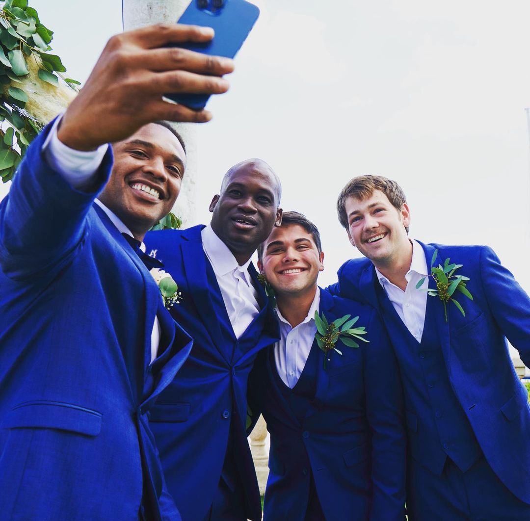 A group of four men in blue suits smiles for a selfie outdoors, with one holding a smartphone to capture the moment.