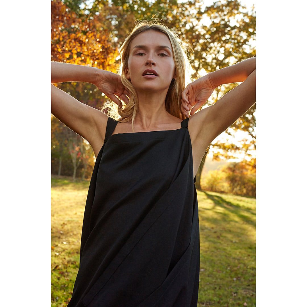 Portrait of a blond woman in a black sundress standing in a grassy field, looking at the camera with trees in the background.