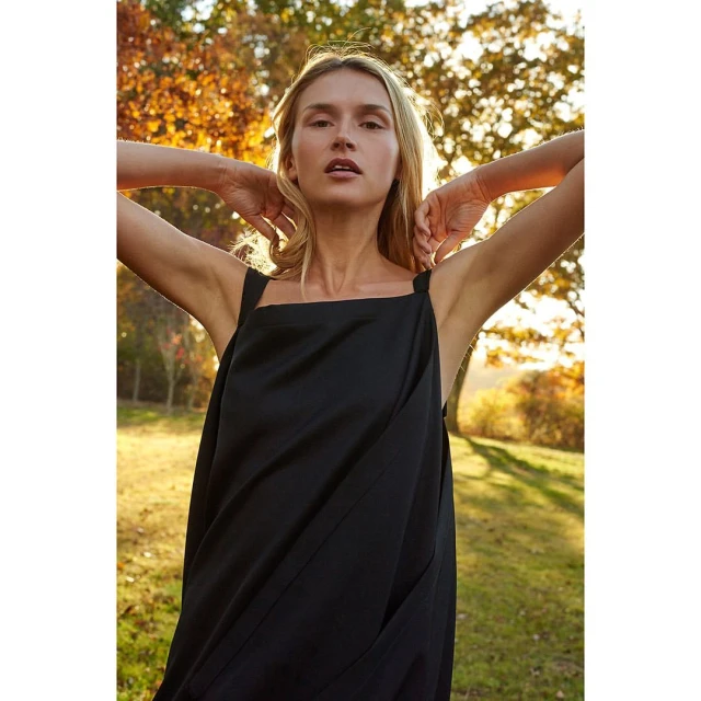 Portrait of a blond woman in a black sundress standing in a grassy field, looking at the camera with trees in the background.