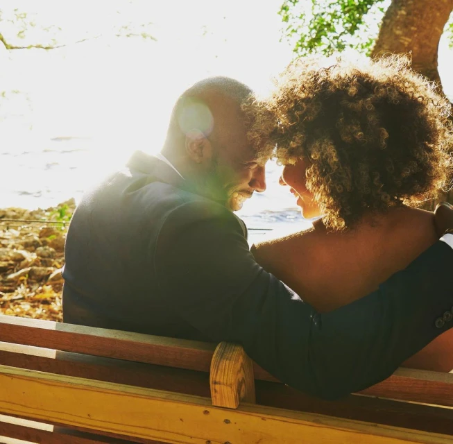 Romantic moment: a dark-suited man embracing a woman with curly hair on a wooden bench under a tree, with sunlight filtering through.