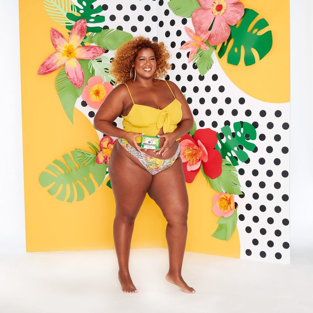 Attractive plus-size Black woman posing with skincare in a studio. She wears a yellow top and patterned bottoms, with paper flowers in the background.