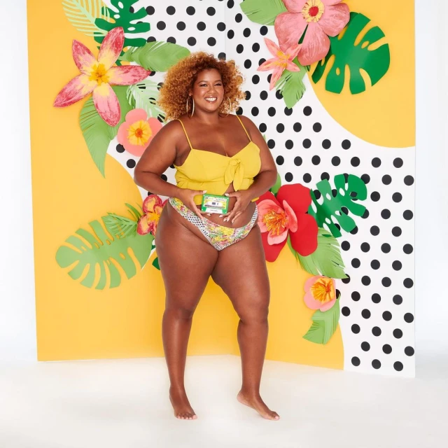 Attractive plus-size Black woman posing with skincare in a studio. She wears a yellow top and patterned bottoms, with paper flowers in the background.