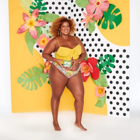 Attractive plus-size Black woman posing with skincare in a studio. She wears a yellow top and patterned bottoms, with paper flowers in the background.