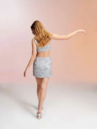Woman in shimmering silver two-piece outfit with matching high-heeled sandals posing in a studio against a peach-toned backdrop.