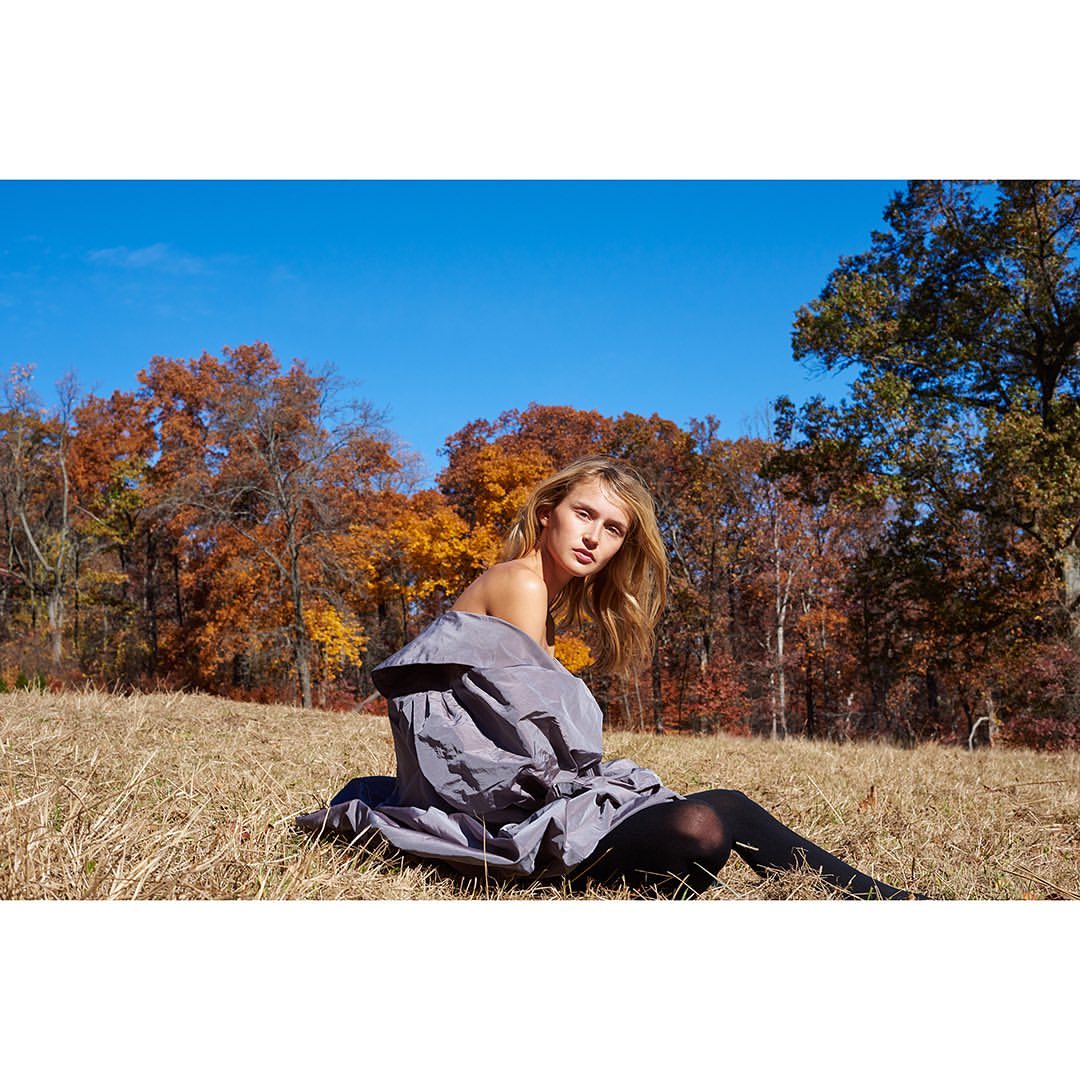 A young woman with long blonde hair poses outdoors in a grey coat and black tights, seated in a grassy field with fall foliage.