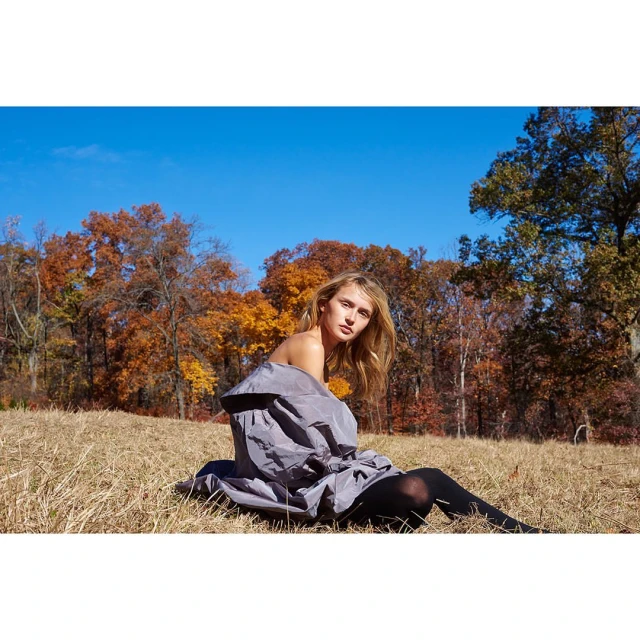 A young woman with long blonde hair poses outdoors in a grey coat and black tights, seated in a grassy field with fall foliage.