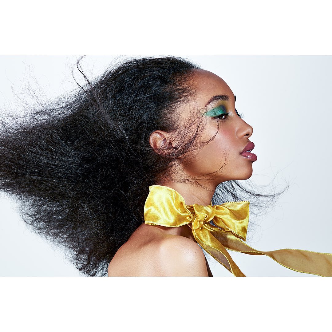 Glamour headshot of a woman with flowing hair and dramatic green eyeshadow, accented by a large golden bow around her neck.