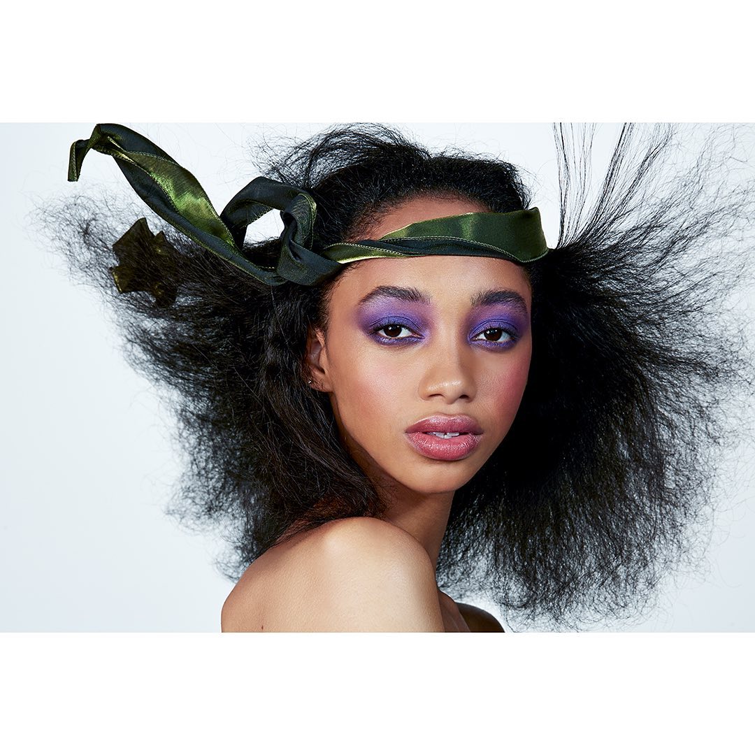 Beauty shot of a woman with a green ribbon head wrap, purple eyeshadow, and dark hair against a clean white backdrop in a studio setting.
