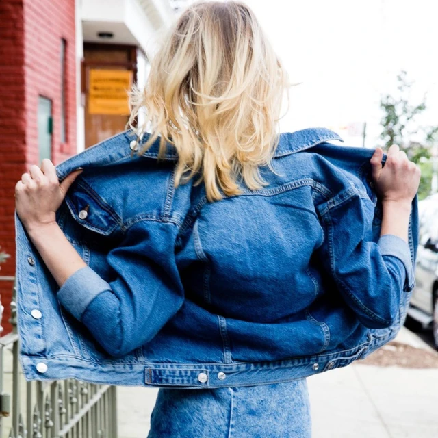 Blonde woman in a denim jacket standing on a sidewalk with a brick building and cars in the background, shot from the rear.