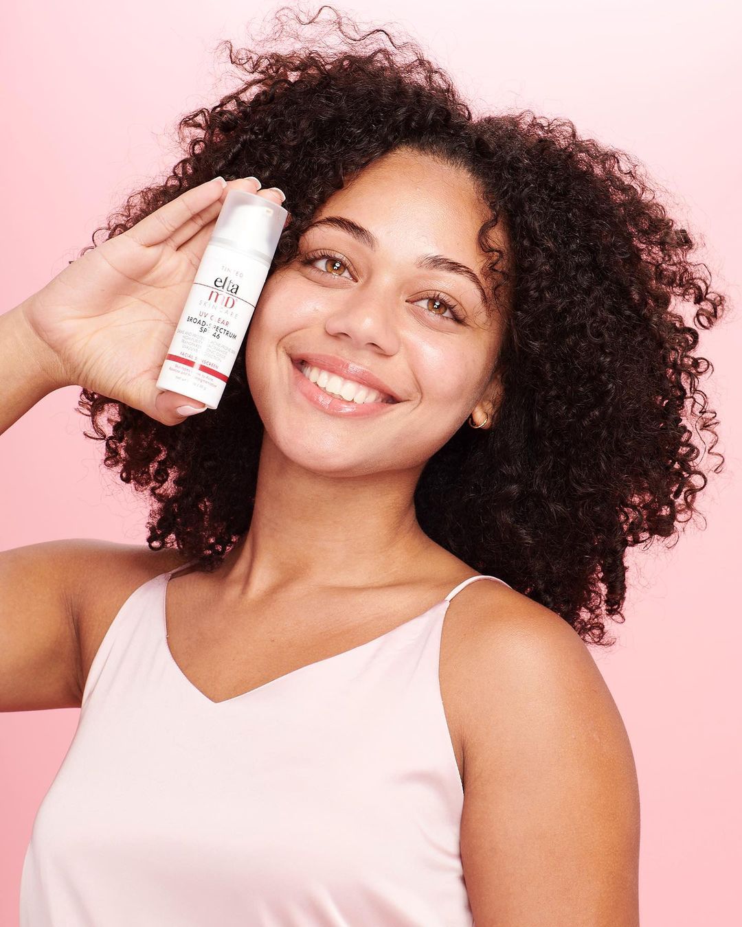 Smiling woman with curly hair holding skin care product in studio with pink backdrop, promoting the brand in beauty advertisement.