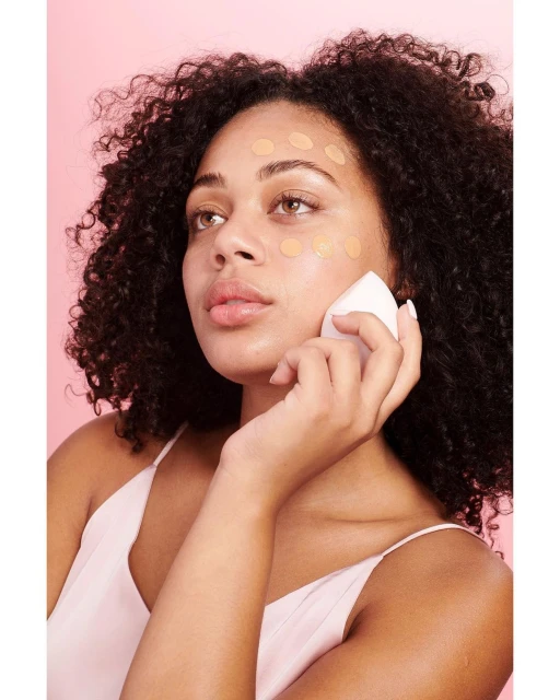 Beauty shot of a woman applying dots of liquid foundation to her face with a white cosmetic sponge on a pink background.
