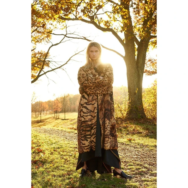 Woman in an animal-print coat stands in a field during golden hour with tall trees and a vibrant sunset illuminating the background.