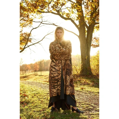 Woman in an animal-print coat stands in a field during golden hour with tall trees and a vibrant sunset illuminating the background.
