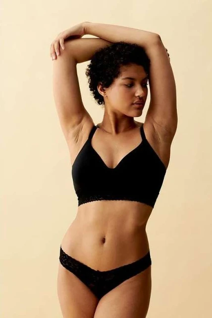 Woman in black bra and underwear poses against a cream background in a studio shot. Her arms are raised over her head.