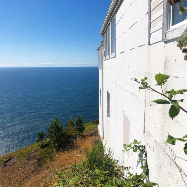 Coastal view of a white building overlooking a blue ocean under a clear sky, with greenery on the hillside, showcasing a natural landscape.