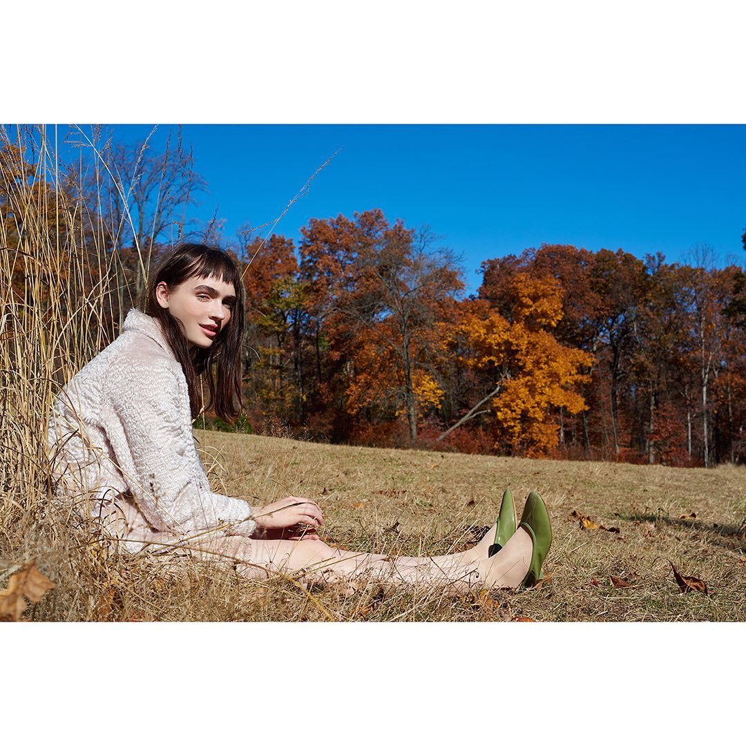 Adult female model with dark hair poses while sitting in a grassy field in front of trees with autumn foliage and a clear blue sky.