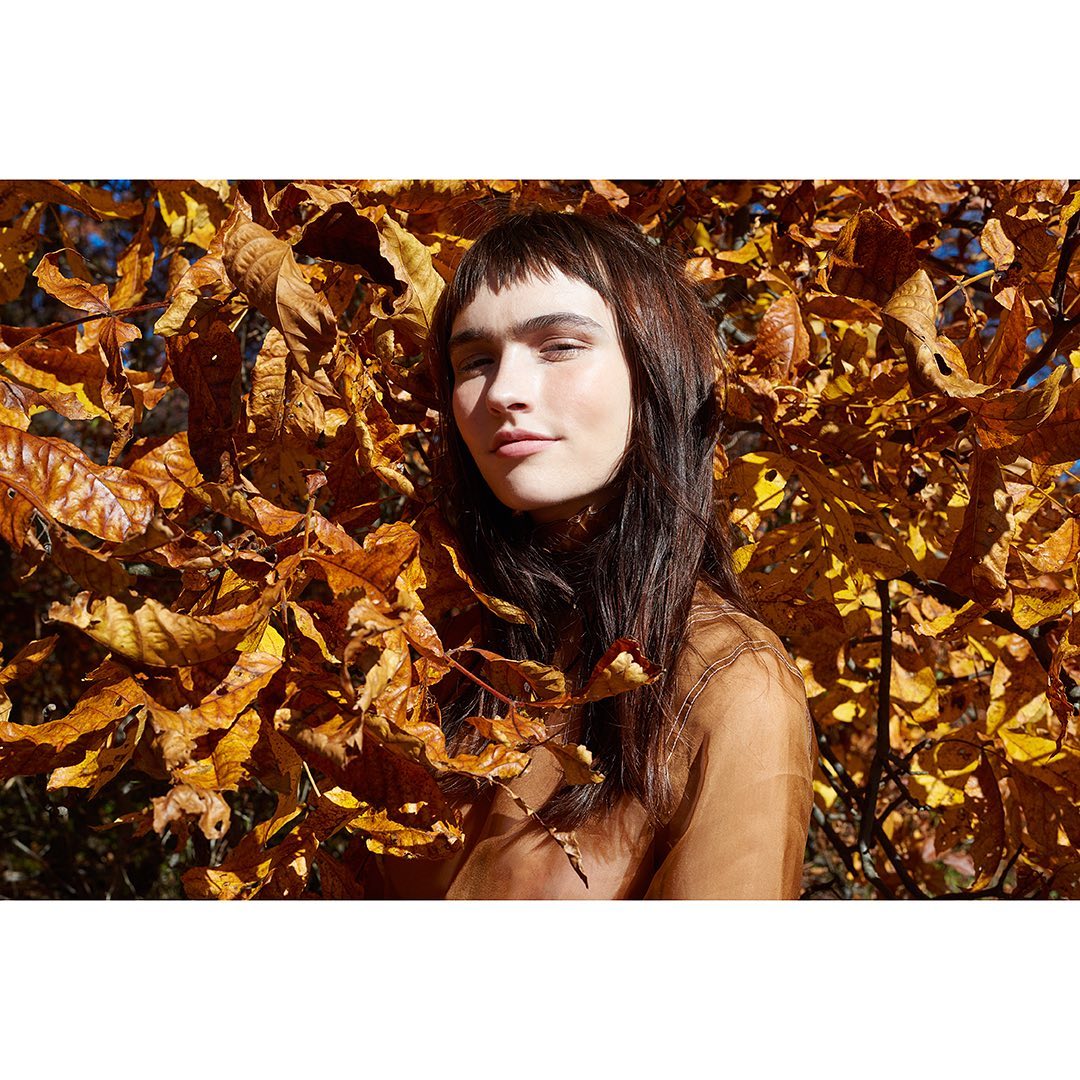 Portrait of a young woman with dark hair and bangs, posing outdoors among a backdrop of golden and brown autumn leaves in natural sunlight.