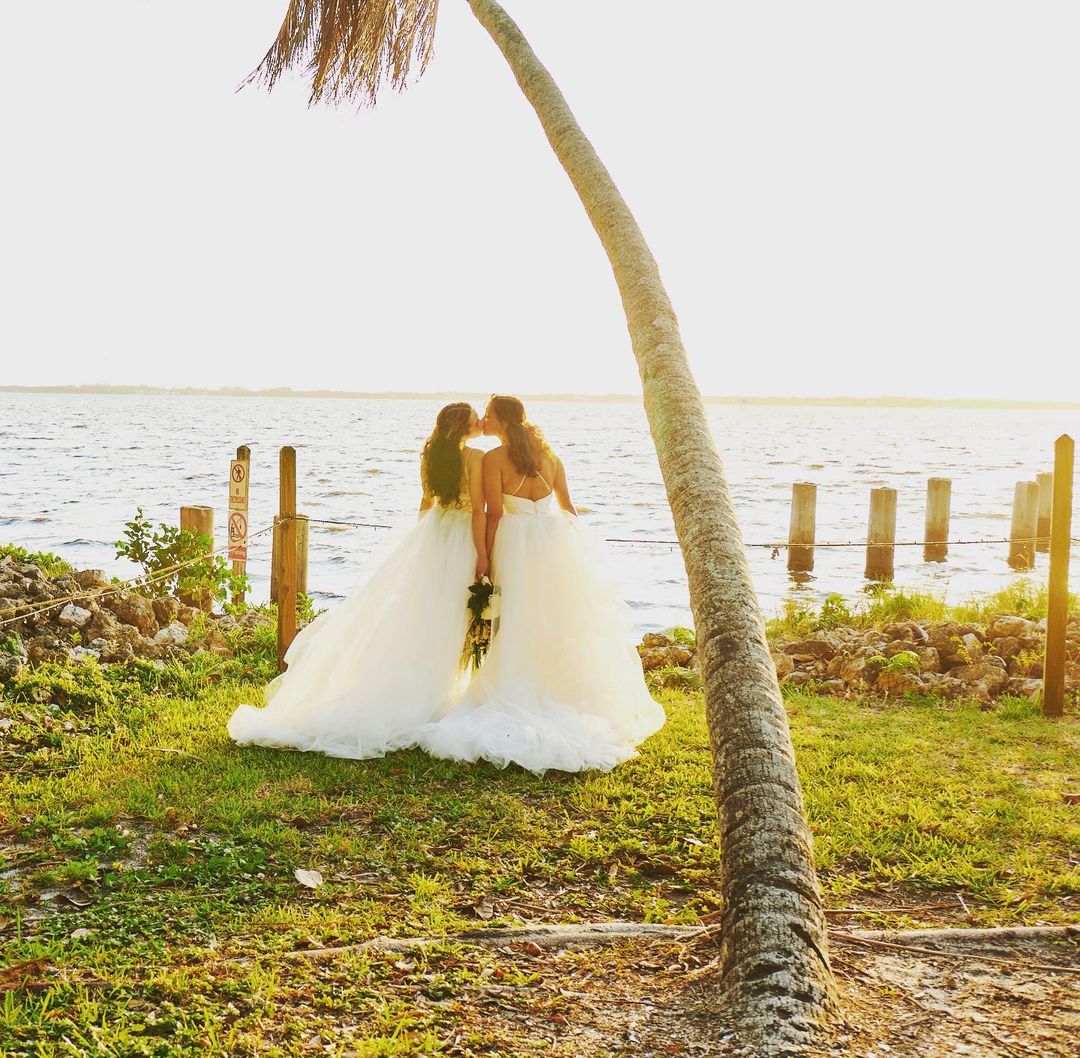 Two brides in white gowns share a kiss near the water at golden hour, with a leaning palm tree framing the idyllic scene.