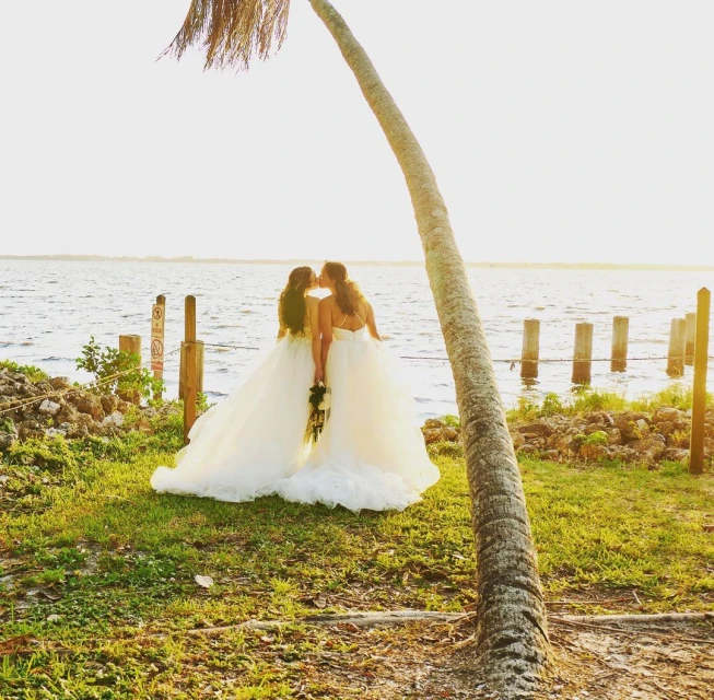 Two brides in white gowns share a kiss near the water at golden hour, with a leaning palm tree framing the idyllic scene.