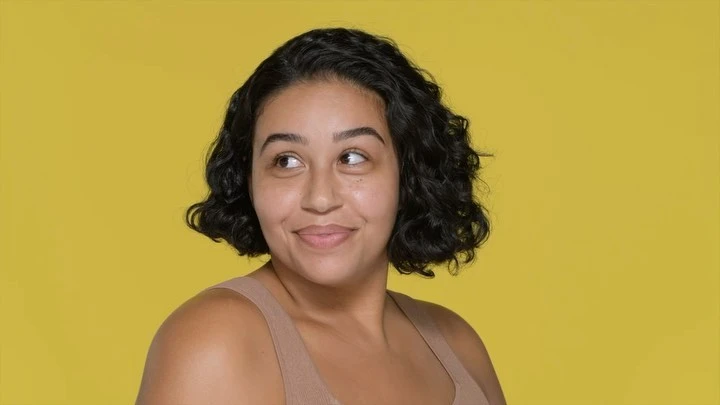 Smiling adult woman with short curly black hair and tan skin, wearing a tan tank top, poses in front of a yellow background.