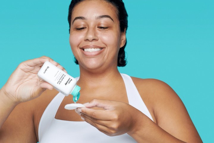 Woman applying skincare product to cotton pad against a turquoise background, demonstrating beauty routine for healthy skin.