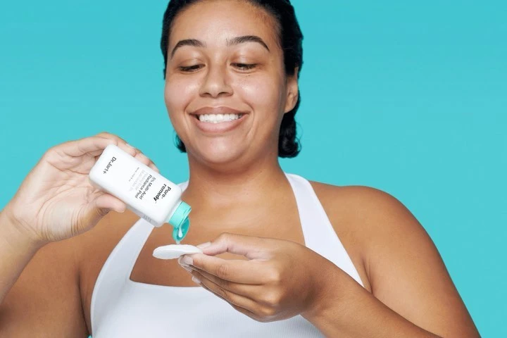 Woman applying skincare product to cotton pad against a turquoise background, demonstrating beauty routine for healthy skin.