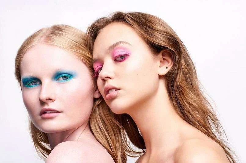 Two women pose in a close-up shot with bright blue and pink eyeshadow makeup against a white background in a beauty studio setting.