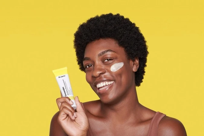 A smiling black woman applies skincare, holding a Ceramidim moisturizing cream tube against a bright yellow studio backdrop.